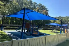 Thumbnail for Children's playground with blue shade structures, slides, and climbing equipment surrounded by trees and white fence.