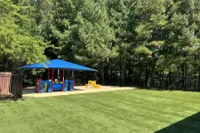 Thumbnail for Children's playground with blue shade cover, colorful play structures, and green grassy area surrounded by trees.