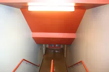 Thumbnail for Indoor stairwell with orange handrails and ceiling leading down to double doors at the bottom.