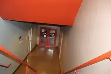 Thumbnail for View down a hallway with red double doors and red handrails on both sides under an orange ceiling overhang.