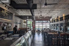 Thumbnail for Interior of a modern cafe with high ceilings, wooden tables, industrial decor, and a glass display counter.