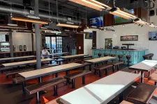 Thumbnail for Modern industrial-style cafeteria with long tables, bench seating, hanging lights, and a bar area in the background