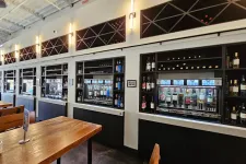 Thumbnail for Modern wine tasting room with wooden tables and automated wine dispensers against white walls.