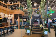 Thumbnail for Modern indoor restaurant with wooden tables, hanging lights, plants, staircase, and arcade games in the background