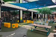 Thumbnail for Outdoor seating area with picnic tables, yellow recycling bins, shade sails, and string lights in a commercial space.