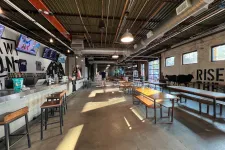 Thumbnail for Spacious brewery tasting room with bar stools, wooden tables, industrial ceiling, and wall art of cows and text.