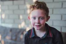 Thumbnail for Young boy with curly red hair wearing a black Dayco Systems shirt outdoors near a brick wall.