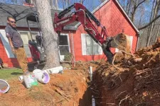 Thumbnail for Excavator digging trench for white PVC pipe installation near red house in a wooded area.