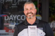 Thumbnail for Smiling man in black Dayco Systems shirt holding fan of company brochures outside business entrance.