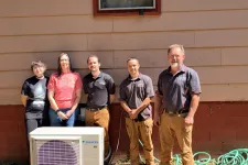 Thumbnail for Five people standing outdoors behind a Daikin air conditioning unit next to a red and beige building wall.