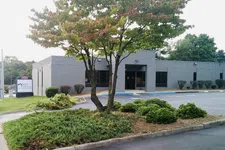 Thumbnail for Gray office building with landscaped greenery and parking lot under a partly cloudy sky.