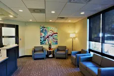 Thumbnail for Modern waiting room with gray chairs, colorful abstract painting, floor lamp, and large windows with dark blinds.