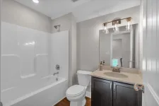 Thumbnail for Modern bathroom with white bathtub, toilet, dark wood vanity, and mirror under bright light fixtures