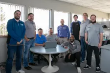Thumbnail for Group of nine diverse men posing around a table with a laptop in a bright office space with large windows.