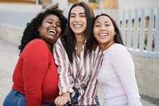 Thumbnail for Three diverse young women smiling and hugging outdoors near a white fence, enjoying a sunny day together.