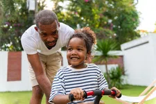 Thumbnail for Father supports smiling daughter riding a red tricycle in a sunny backyard with greenery and flowers.
