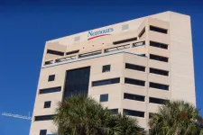 Thumbnail for Modern beige office building with Nemours logo atop under clear blue sky and palm trees in foreground