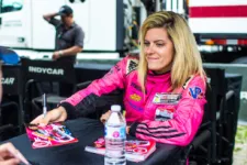 Thumbnail for Female race car driver in pink suit signing autographs for a young fan at an IndyCar event.