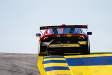 Thumbnail for Rear view of a Lamborghini race car speeding on a race track with yellow and blue curbs under a clear sky.