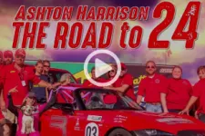 Thumbnail for Group of people posing with a red race car under the title Ashton Harrison The Road to 24 with a sunset sky.