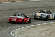Thumbnail for Two Mazda MX-5 race cars, red and gray, competing on a curved asphalt race track in a motorsport event.