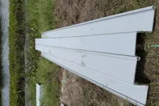 Thumbnail for White metal roofing panels stacked on grassy ground near a water body and plants.