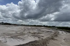 Thumbnail for Wide sandy construction site under a dramatic cloudy sky with distant houses and greenery on horizon.