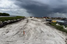 Thumbnail for Construction site with dirt road and equipment under dark stormy clouds near waterfront and vegetation.