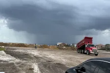 Thumbnail for Red dump truck unloading soil on construction site under dark rainy clouds with worker and houses in background.