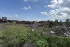 Thumbnail for Cleared land with piles of debris and construction equipment under a partly cloudy blue sky.