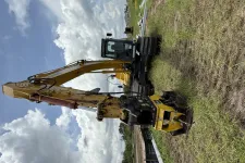 Thumbnail for Yellow excavator with hydraulic arm and metal attachments parked on grassy field under cloudy sky.