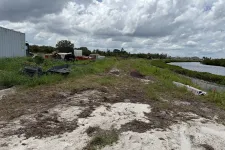 Thumbnail for Dirt pathway with tire tracks beside a body of water under cloudy sky and scattered construction materials nearby