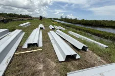 Thumbnail for Metal sheet piles laid out on grass near a canal under a cloudy sky at a construction site.