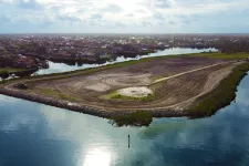 Thumbnail for Aerial view of a large cleared land area surrounded by water with residential houses in the background under a cloudy sky.