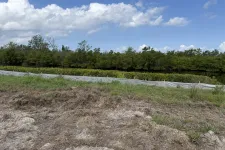 Thumbnail for Cleared land with dirt foreground, green grassy area, white barrier fence, and dense trees under a partly cloudy blue sky.