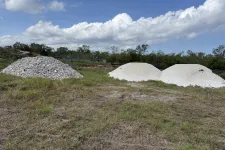 Thumbnail for Three piles of gravel and crushed stones on grassy land under a partly cloudy blue sky near trees.