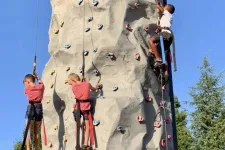 Thumbnail for Three children climbing a tall outdoor rock climbing wall with colorful holds on a sunny day.