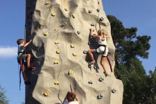 Thumbnail for Three children climbing a large outdoor rock climbing wall with safety harnesses on a clear sunny day.