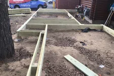 Thumbnail for Wooden frame structure for a deck or small building under construction on dirt ground near a log cabin.