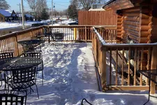 Thumbnail for Snow-covered outdoor patio with metal tables and chairs next to a wooden log cabin under clear blue sky.