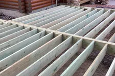 Thumbnail for Wooden deck frame under construction next to a log cabin with stone porch columns outdoors.