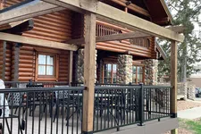 Thumbnail for Log cabin porch with wooden beams, stone pillars, black metal railing, and outdoor seating under trees.