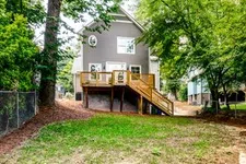 Thumbnail for Backyard view of a gray two-story house with wooden deck and stairs surrounded by trees and grass.