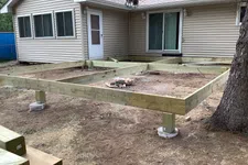 Thumbnail for Wooden deck frame construction supported by concrete piers next to a beige house with windows and sliding door.