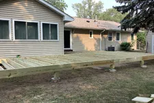 Thumbnail for Wooden deck framework under construction beside a single-story house in a backyard with trees and grass.