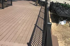 Thumbnail for Empty brown wooden deck with black metal railing and stacked outdoor chairs beside trees in sunny weather.