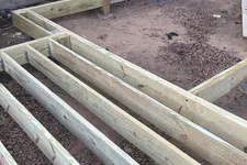 Thumbnail for Wooden frame foundation construction for a deck or small building next to a log cabin on gravel and dirt.