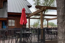 Thumbnail for Outdoor seating area with black metal chairs and tables, red closed umbrella, wooden pergola, and log cabin exterior.