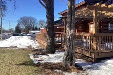 Thumbnail for Log cabin with wooden deck surrounded by melting snow and two large trees on a clear sunny day.