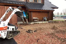 Thumbnail for Mini excavator with auger attachment clearing rocky soil beside wooden cabin building under construction.